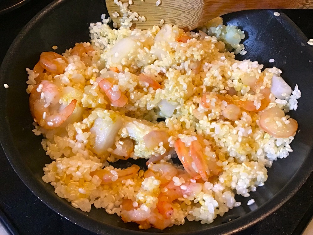 フライパンにオリーブオイルを入れ、研いだ米を炒める。米が少し透き通ってきたら、冷凍のシーフードミックスを入れてさらに炒める。