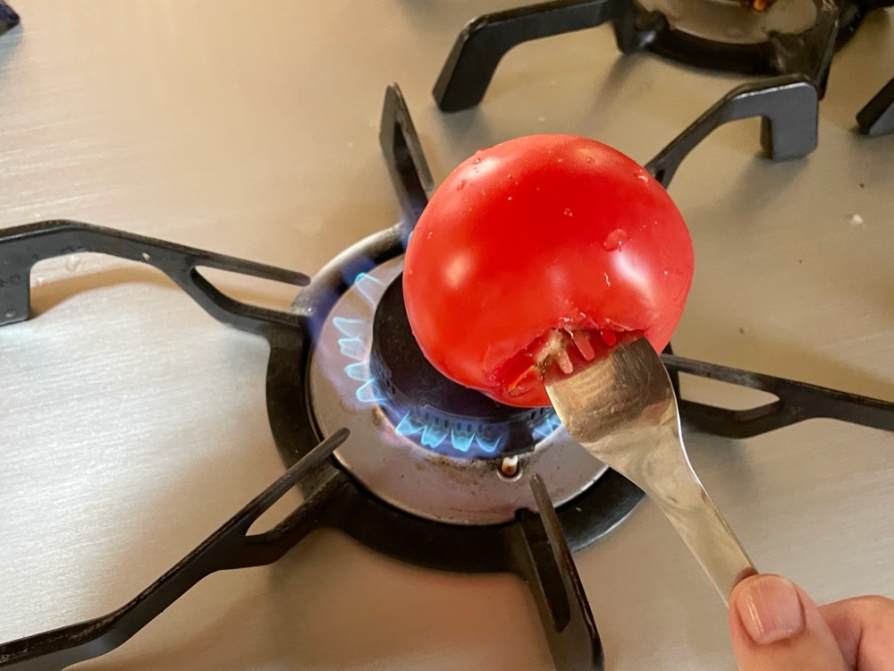 ヘタを取ったところにフォークを刺して直火であぶり、皮がめくれてきたら水に取って皮を剥く。